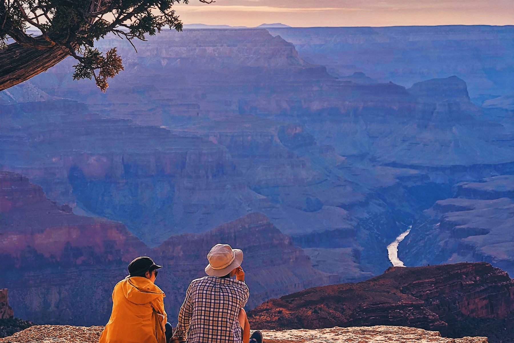 A couple sits at a Grand Canyon overlook at sunset, taking in the view of the canyon and the Colorado River below